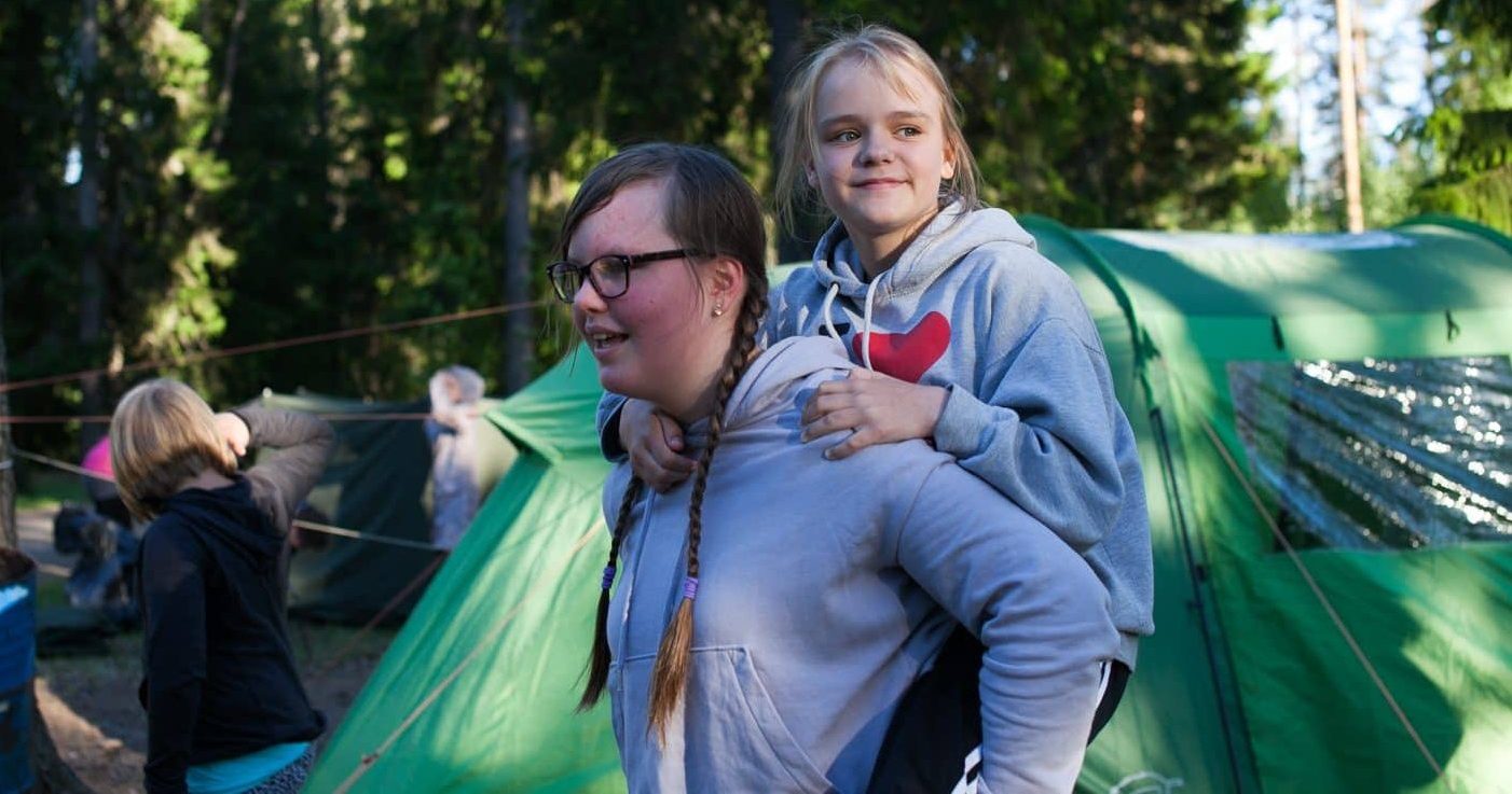Lapsi on toisen lapsen reppuselässä telttaleirillä.